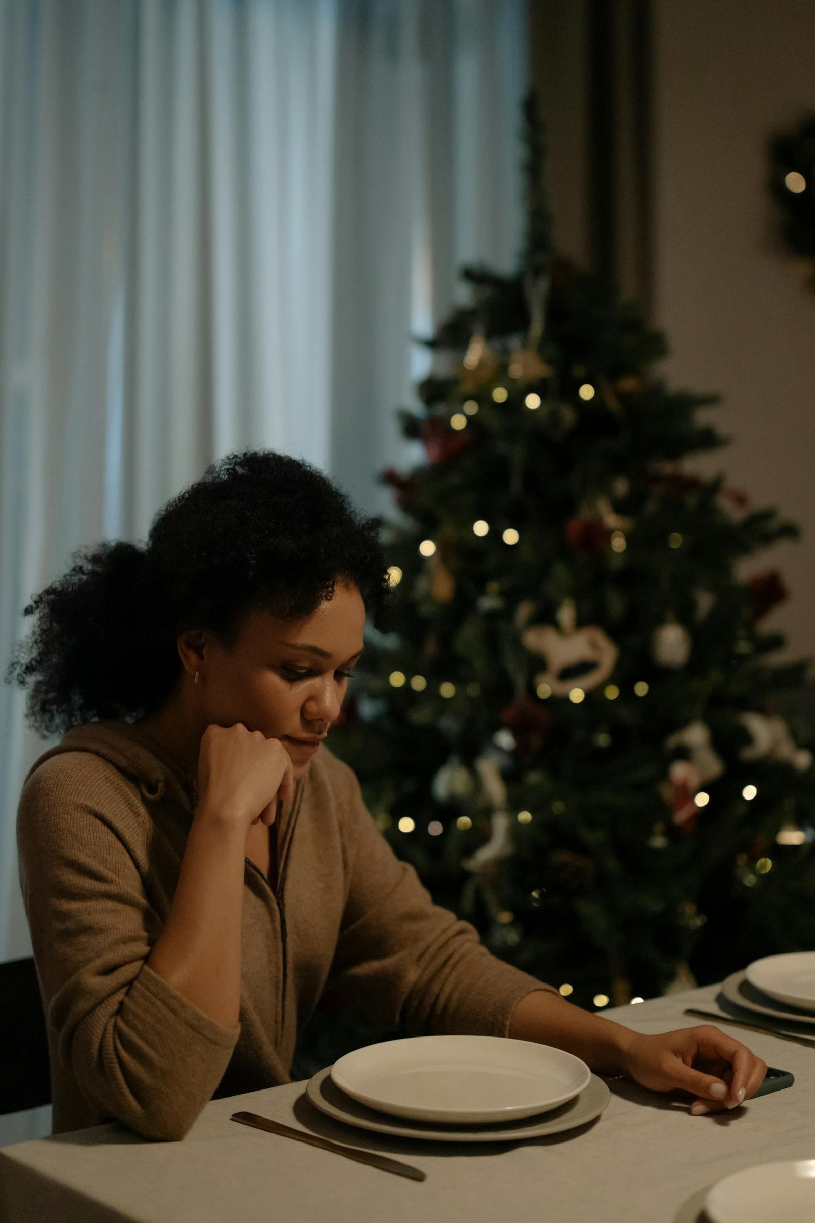 A solitary woman sitting at a dining table beside a decorated Christmas tree.